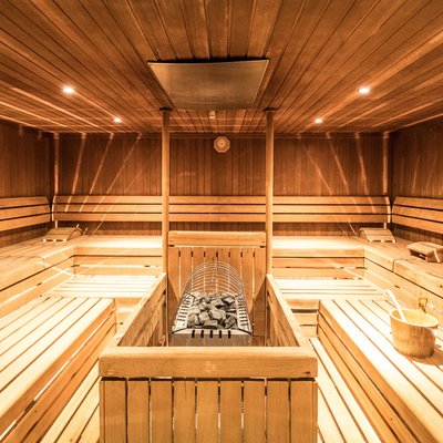 Auf dem Bild ist der Innenraum einer Sauna gezeigt. Der Raum ist vollständig mit holzverkleideten Wänden, Decke und Sitzbänken ausgestattet, was den typischen Look einer finnischen Sauna vermittelt. Entlang beider Seiten des Raums befinden sich mehrere Holzbänke in drei Sitzebenen, die übereinander angeordnet sind und ausreichend Platz für mehrere Personen bieten. In der Raummitte steht ein Saunaofen mit aufgelegten Saunasteinen, der die Wärme erzeugt. Daneben steht ein hölzerner Sauna‑Wasserkübel mit einer Schöpfkelle, mit dem Wasser auf die Steine gegeben werden kann, um Dampf zu erzeugen. An der Rückwand ist ein rundes Thermometer oder Hygrometer angebracht, um Temperatur oder Luftfeuchtigkeit abzulesen. Die Beleuchtung ist dezent und gleichmäßig, die Atmosphäre wirkt warm und einladend.