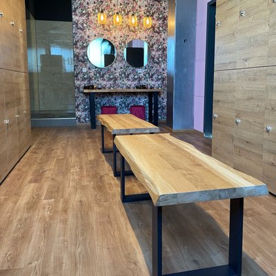 Raumansicht der Damen-Umkleide im Fitness Express Gärtringen mit Holzbänken und Holzschränken. Im Vordergrund stehen zwei lange, hellbraune Holzbänke mit schwarzen Metallgestellen auf einem hellen Holzfußboden. Rechts und links sind hohe Schließfächer aus Holz mit silbernen Griffen angeordnet. An der hinteren Wand ist eine Tischfläche mit zwei runden Spiegeln darüber angebracht. Darüber hängen vier warme Hängeleuchten mit bernsteinfarbenem Licht. Die Rückwand zeigt eine Blüten-/pflanzenartige Tapete, links davon ist ein Dusch- oder Sanitärbereich mit Glas und hellen Fliesen.