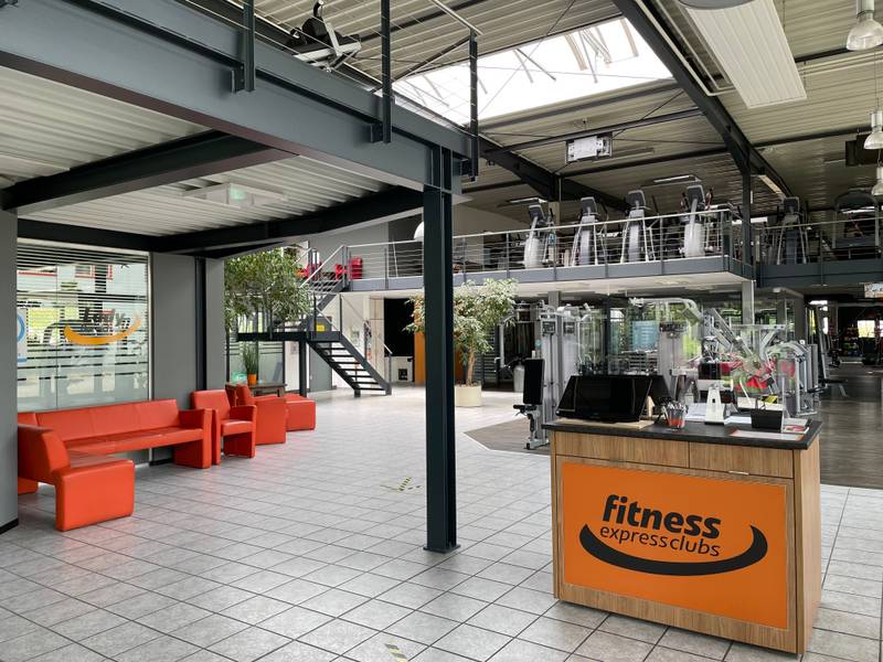 Das Bild zeigt den Eingangs‑ und Empfangsbereich eines Fitnessstudios (Fitness Express Clubs). Im Vordergrund steht ein Empfangstresen mit dem Orange‑schwarzen Logo „fitness expressclubs“. Rechts und links daneben ist ein offener, weitläufiger Raum mit Trainingsgeräten zu sehen, darunter Kraft‑ und Ausdauergeräte auf zwei Ebenen. Links im Bild stehen mehrere orangefarbene Sitzmöbel (Sofa und Sessel) für wartende oder ruhende Personen. Große Fensterelemente und ein Dach mit sichtbaren Stahlträgern lassen viel Licht in den Raum. Insgesamt wirkt der Bereich hell, offen und funktional mit klar erkennbarem Check‑in bzw. Empfang vorne