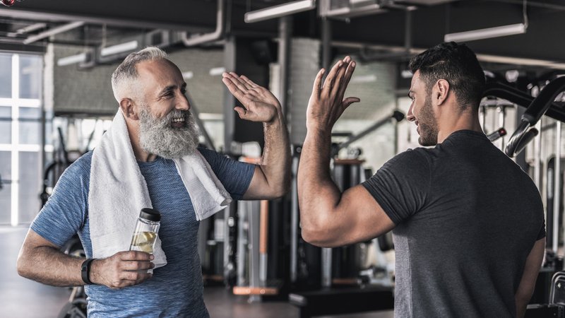 Das Bild zeigt zwei Männer in einem modernen Fitnessstudio mit verschiedenen Trainingsgeräten im Hintergrund. Der Mann links ist älter mit grauem Haar und Bart, trägt ein sportliches T‑Shirt, hat ein weißes Handtuch über der Schulter und hält eine durchsichtige Trinkflasche in der linken Hand. Er lächelt und hebt seine rechte Hand zum High‑Five. Der Mann rechts ist muskulös, trägt ein dunkles Sportshirt und hebt ebenfalls seine rechte Hand zum High‑Five. Beide Personen stehen sich gegenüber und feiern mit dem High‑Five offenbar gemeinsam einen Erfolg oder das Ende eines Trainings.