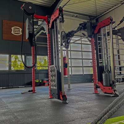 Auf dem Foto ist der Functional‑Trainingsbereich im Fitness Express Böblingen zu sehen. Zentral im Bild steht ein großer Funktional-Trainingsturm aus Metall, das in Schwarz und Rot gehalten ist und verschiedene Trainingsmöglichkeiten bietet, etwa Stationen zum Ziehen, Drücken und Hängen von Fitness‑Zubehör. Am Boden liegt ein dunkler, gummierter Bodenbelag, der typischerweise in Functional‑Bereichen verwendet wird, um Sicherheit und Dämpfung zu gewährleisten.  Im Vordergrund rechts liegen dicke Seile (Battle Ropes), die für Kraft‑ und Ausdauerübungen benutzt werden. Im Hintergrund links sind gestapelte Trainings‑Zusatzgeräte zu erkennen, darunter bunte Sandbags oder Medizinbälle und weitere Kleingeräte, die für funktionelles Training eingesetzt werden können.  Links an der Wand hängt eine Holztafel mit dem Logo „Hammer Strength“, einem bekannten Fitnessgeräte‑Hersteller. Helle Fensterflächen an der rechten Seite lassen Tageslicht in den Raum und schaffen eine gut belichtete, offene Trainingsatmosphäre. Der Raum wirkt geräumig und für vielfältige funktionelle Trainingsformen nutzbar, wie sie im Studio angeboten werden