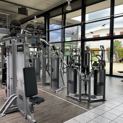 Das Bild zeigt einen hellen, modernen Trainingsbereich im Fitness Express Böblingen mit einer großen Fensterfront die über 2 Stockwerke geht. Der Boden ist dunkelbraun und teils hellgrau. Im Vordergrund stehen mehrere Krafttrainingsgeräte aus Metall mit schwarzen oder dunkelgrauen Polstern, Sitzen, Griffen und Hebeln, die Arme, Beine und Oberkörper trainieren. Zwischen den Geräten ist genügend Platz zum Gehen. Im Hintergrund ist ein zweites Stockwerk mit Geländer zu sehen, das den Raum offen wirken lässt. Von der Decke hängen Lampen, die den Bereich gleichmäßig ausleuchten. Insgesamt wirkt das Studio professionell, ordentlich und modern.