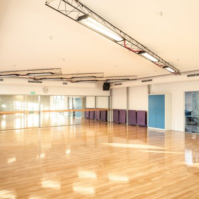 Auf dem Foto ist ein großer, heller Übungsraum in einem Fitnessstudio zu sehen. Der Raum hat einen glatten, hellen Holz­boden und eine hohe Decke mit mehreren langen, rechteckigen Decken­leuchten. Entlang einer Wand befindet sich ein großer Spiegel, der den Raum optisch erweitert. Vor dem Spiegel stehen an einer Seite des Zimmers mehrere gestapelte, rechteckige Gymnastikmatten in dunkler Farbe und ein Regal mit weiteren Fitness­geräten. Rechts an der Wand sind Schränke und durch eine Glas­tür ist ein weiterer Raum erkennbar. Das Tages­licht fällt durch Fenster und schafft eine freundliche, offene Atmosphäre. Im Raum sind keine Personen zu sehen.