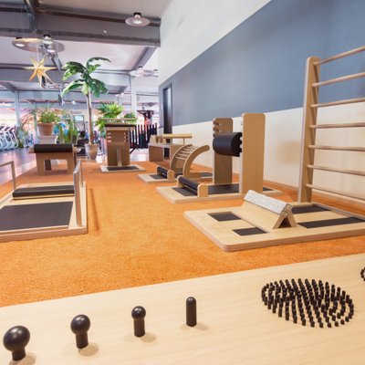 Das Bild zeigt einen hellen Trainingsbereich mit Holzgeräten auf orangefarbenem Boden für Balance, Dehnung und funktionelle Übungen. Im Hintergrund sind große Fenster, Cardio-Geräte und grüne Pflanzen zu sehen. Die Decke ist hell mit Lampen, und der Raum wirkt einladend, offen und ruhig.